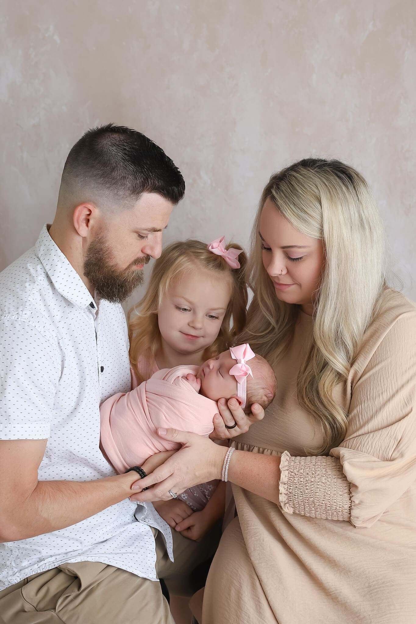 Happy mom, dad and big sister look down at sleeping newborn girl in dad's hands in a pink swaddle