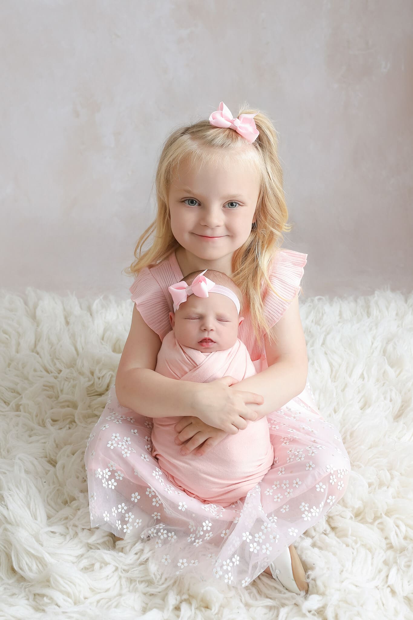 A happy toddler in a pink dress sits cross legged with her newborn baby sister sleeping upright in a pink swaddle in her lap after meeting Nannies In Kansas City