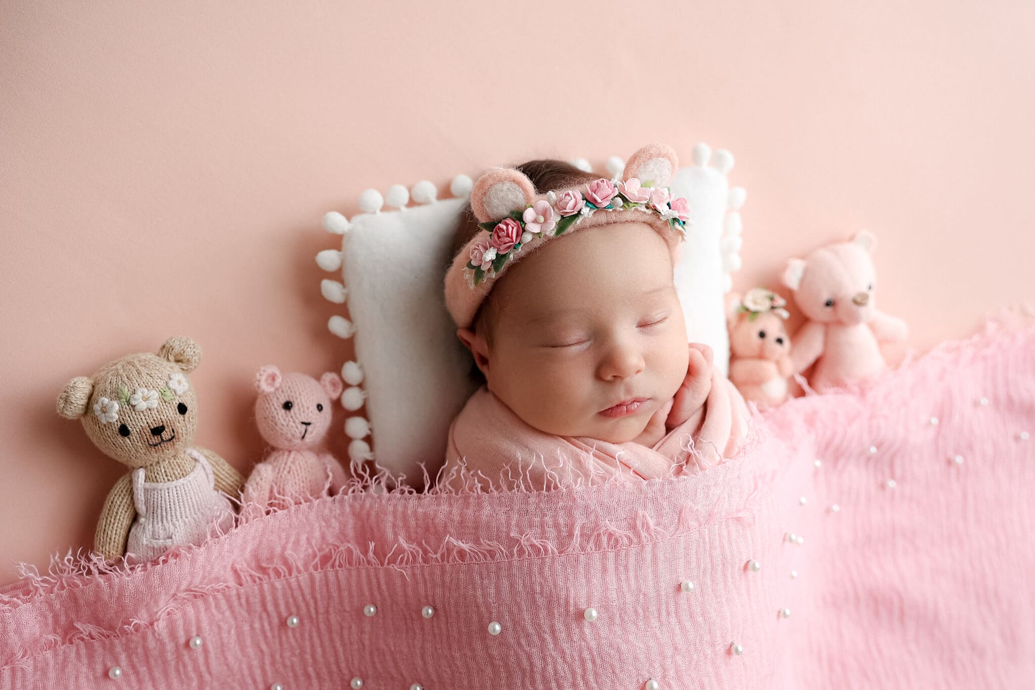 A newborn baby girl sleeps under a pink blanket with a matching teddy bear headband and surrounded by knit teddies after meeting Nannies In Kansas City