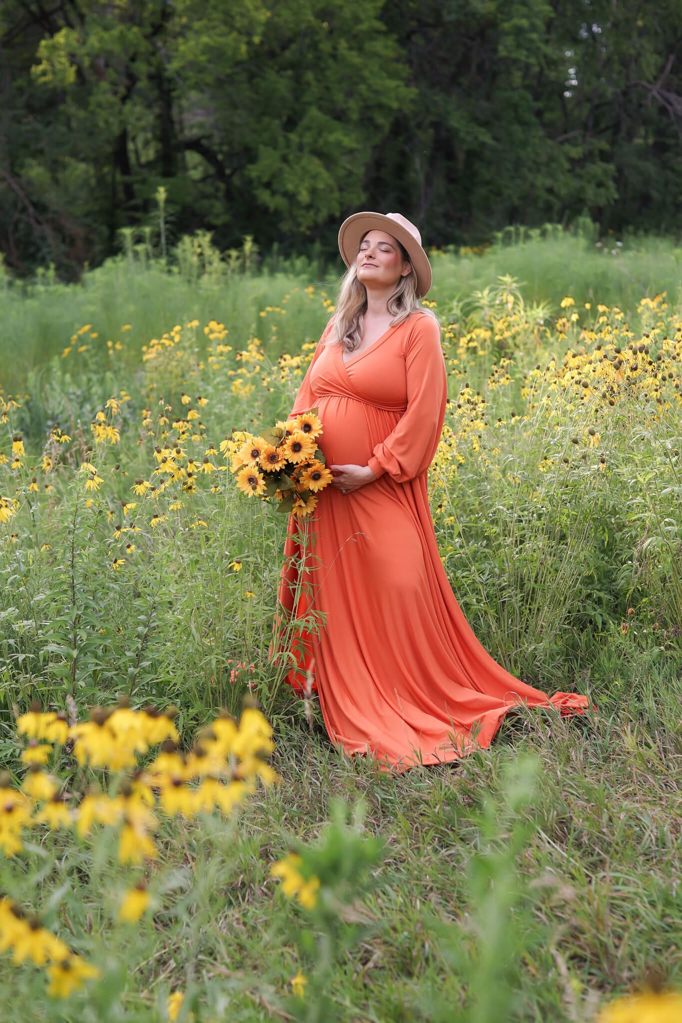 A happy expectant woman in an orange maternity gown stands in a field of yellow wildflowers with eyes closed with a large brown hat and bouquet of sunflowers