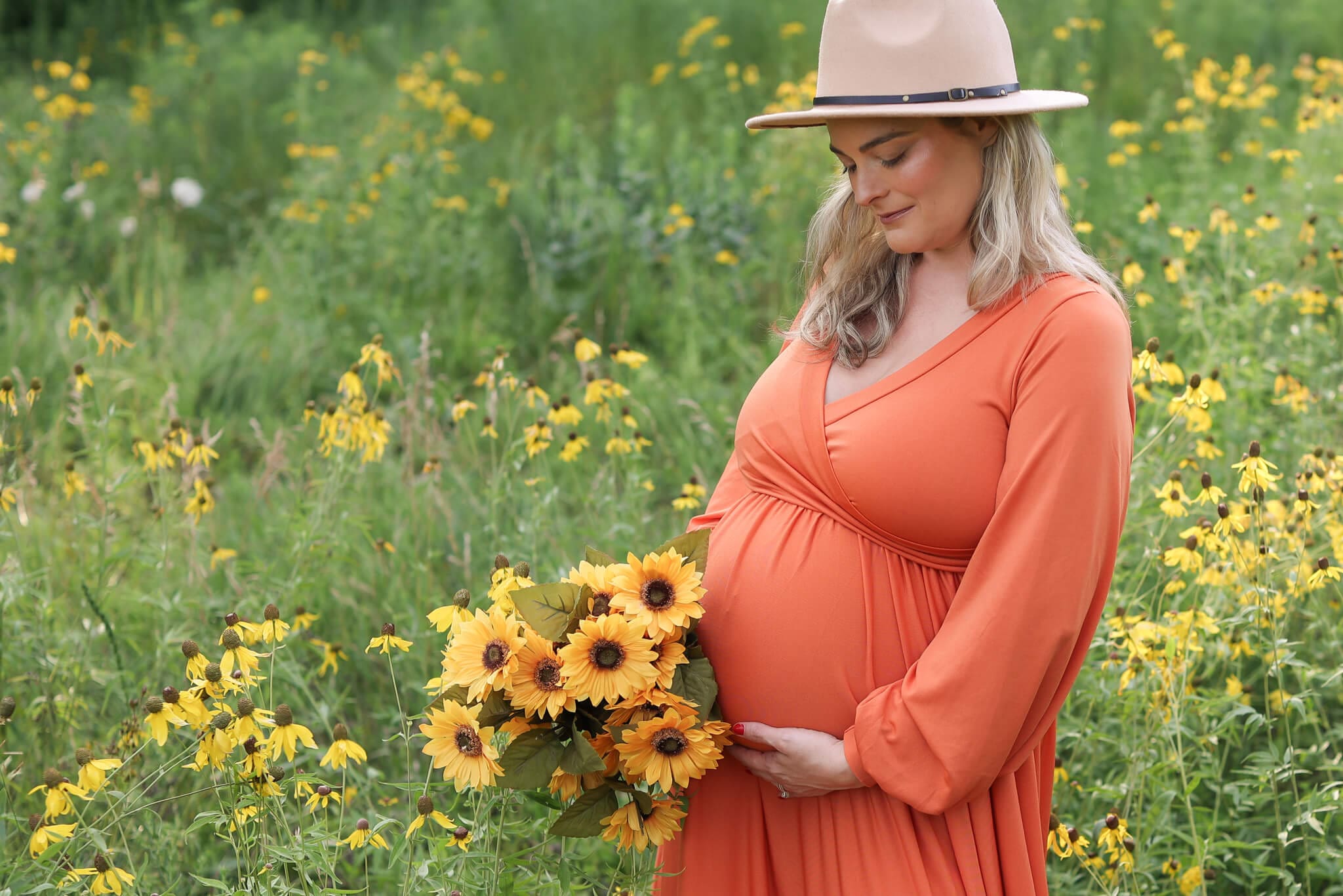 After enjoying a great Prenatal Massage In Kansas City, a pregnant woman smiles down to her bump while standing in a field of wildflowers holding a matching bouquet in an orange gown