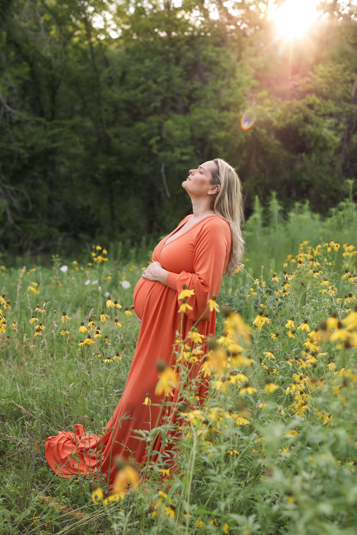 A mother to be in a long orange maternity gown basks in the sun in a field of yellow wildflowers with a hand on her bump after a Prenatal Massage In Kansas City